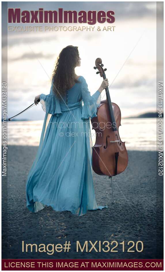 Woman in blue dress standing on a sea shore holding a cello Rear view of her back in dramatic sunrise