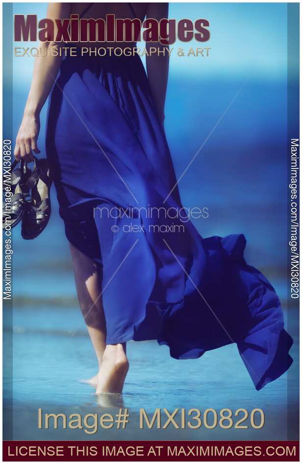 Woman in blue summer dress blowing in the wind walking barefoot on ocean beach sand