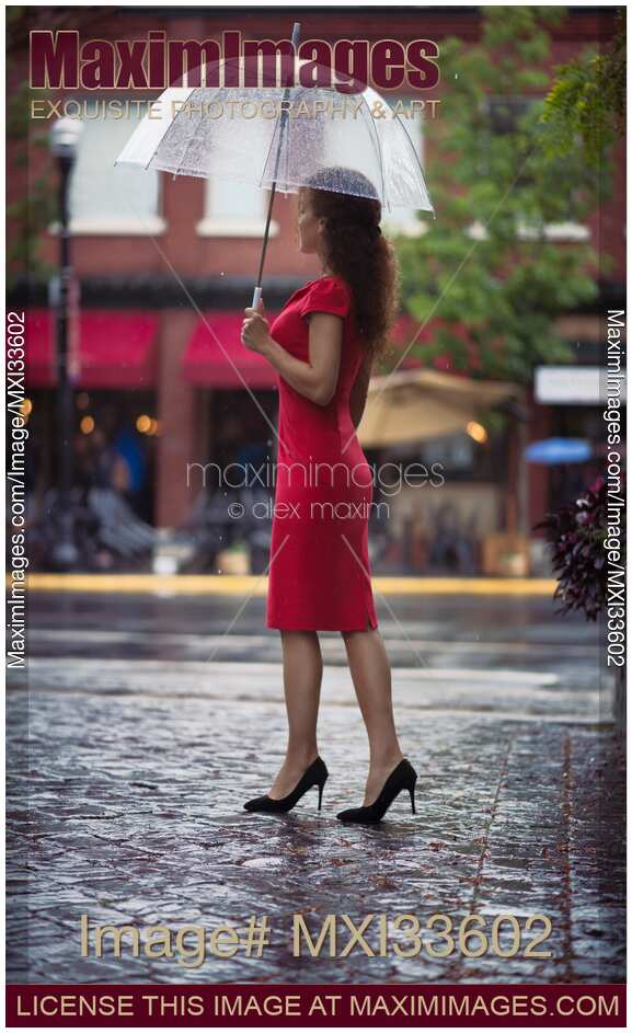 Woman in elegant red dress waiting for someone on a city street with an umbrella under the rain