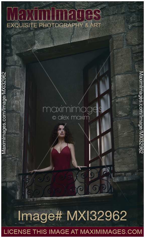 Woman in red dress standing in an open window in a French stone house looking outside at night