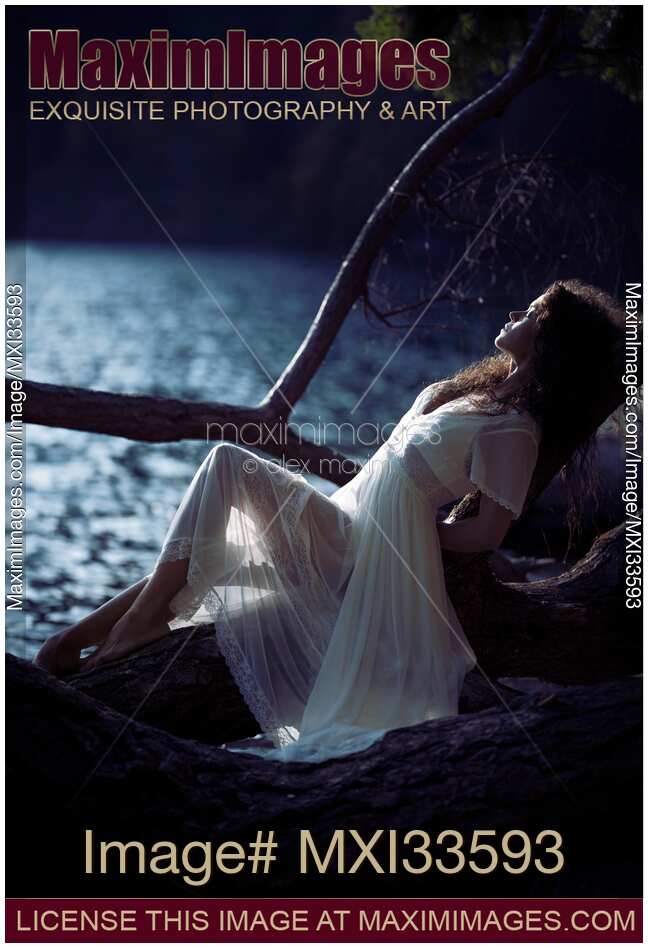 Woman in sheer white lace dress reclining against a tree by the lake in the nature at night in moonlight