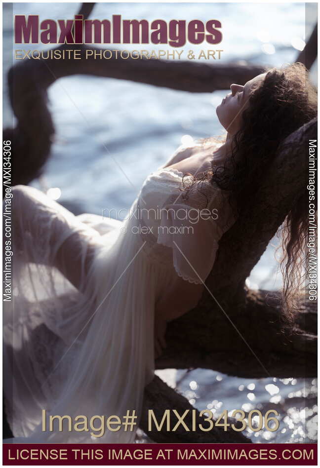 Woman in sheer white lace gown reclining relaxed against a tree by the lake in the nature