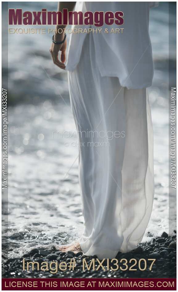 Woman in white summer clothes standing on a pebble shore with fresh cool sea water washing over her bare feet