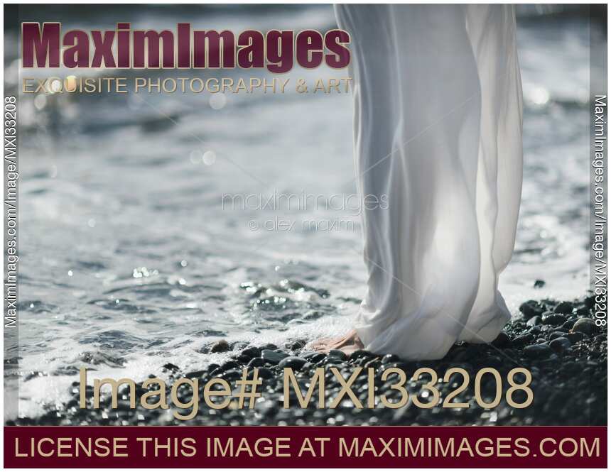Woman in white summer pants standing barefoot on a pebbled ocean beach shore, cool sea water washing over her bare feet