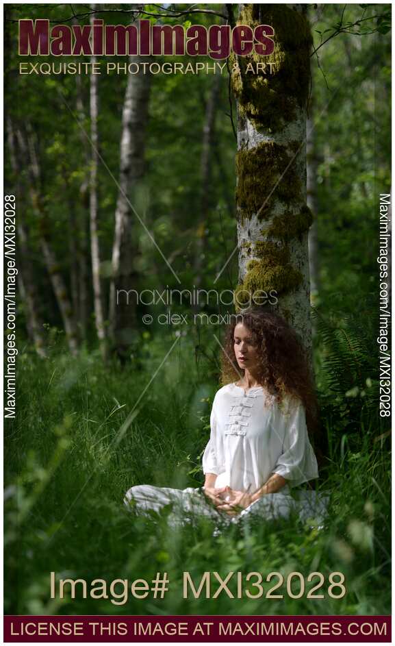 Woman practicing sitting meditation leaning against a birch tree in green forest