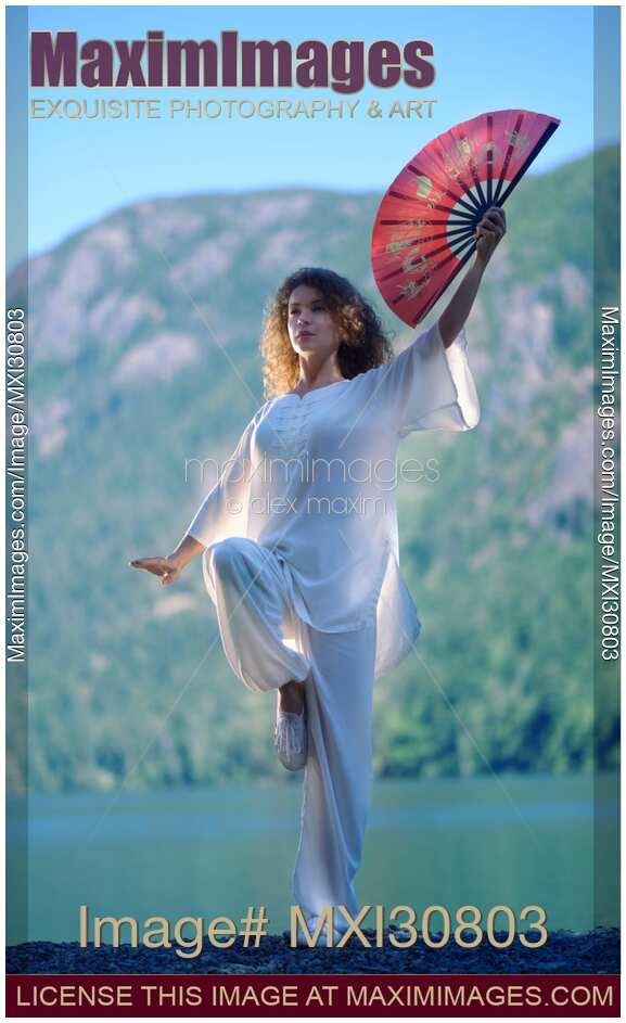 Woman practicing Tai Chi crane stance with a fan at the mountain lake shore in the morning
