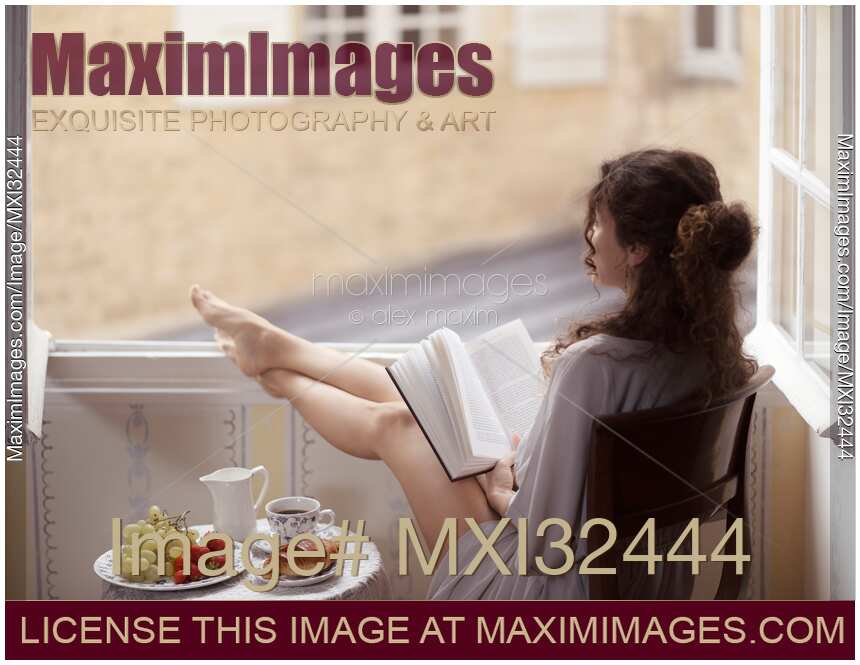 Woman reading a book with her legs on an open window frame and a plate with coffee and croissant