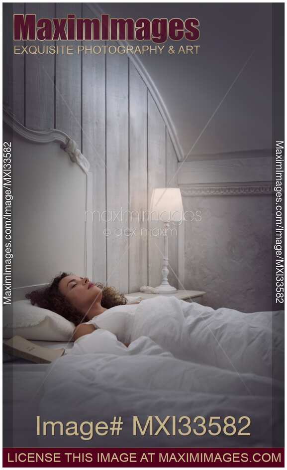 Woman sleeping in bed alone in white vintage room with a book and a lamp beside her