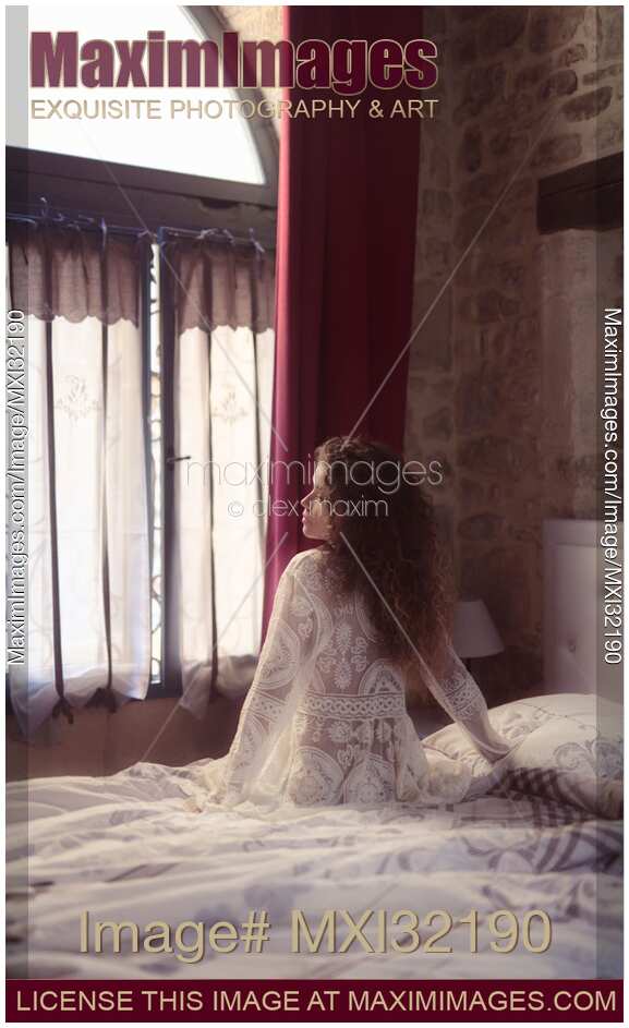 Woman thoughtfully sitting on the side of a bed in French bedroom in white nightgown