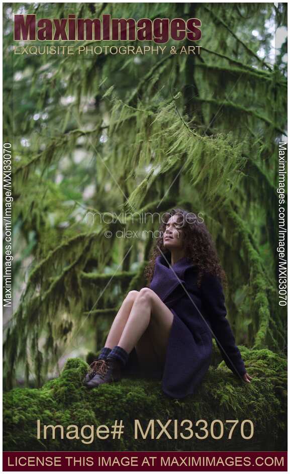 Woman with dreamy expression sitting on mossy tree trunk in purple coat with bare legs in moody green forest nature scenery