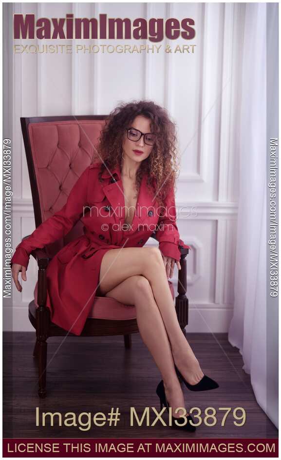 Woman with sexy bared legs in red trench coat and glasses sitting in a chair with thoughtful look