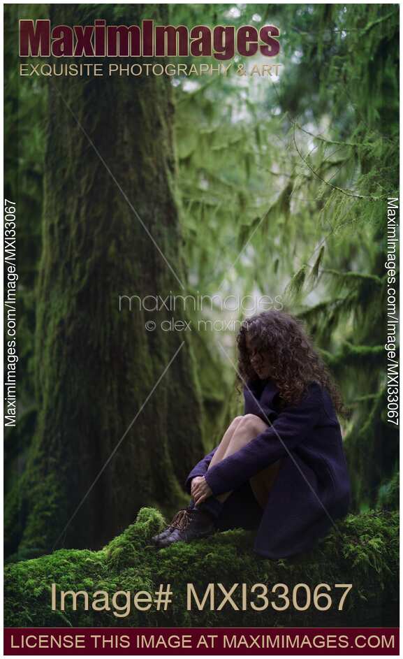 Woman with thoughtful contemplative expression sitting on mossy tree trunk in deep green forest with hands around her legs