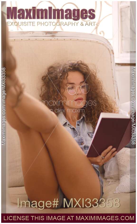 Young carefree woman in glasses and tiny shorts with sexy long legs reading a book in a sensual pose by a window