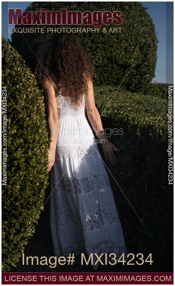 Young lady in white summer dress walking into a green garden disappearing behind the bushes