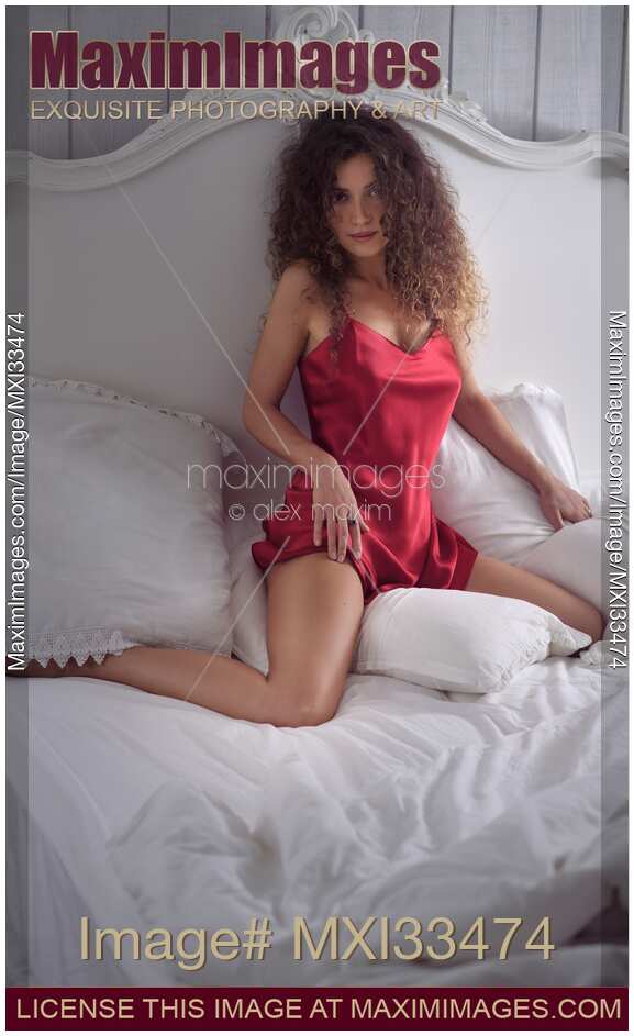 Young pretty woman in red sexy silk nightgown sitting on a bed with an innocent teasing expression