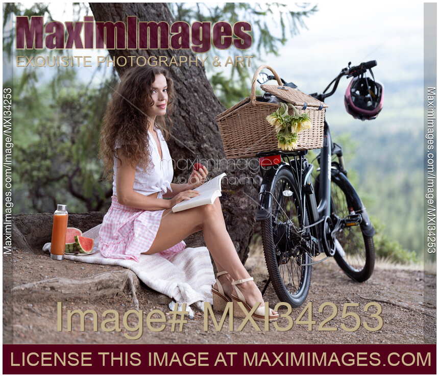 Young pretty woman sitting on a blanket with book and bike enjoying a little picnic in summer countryside