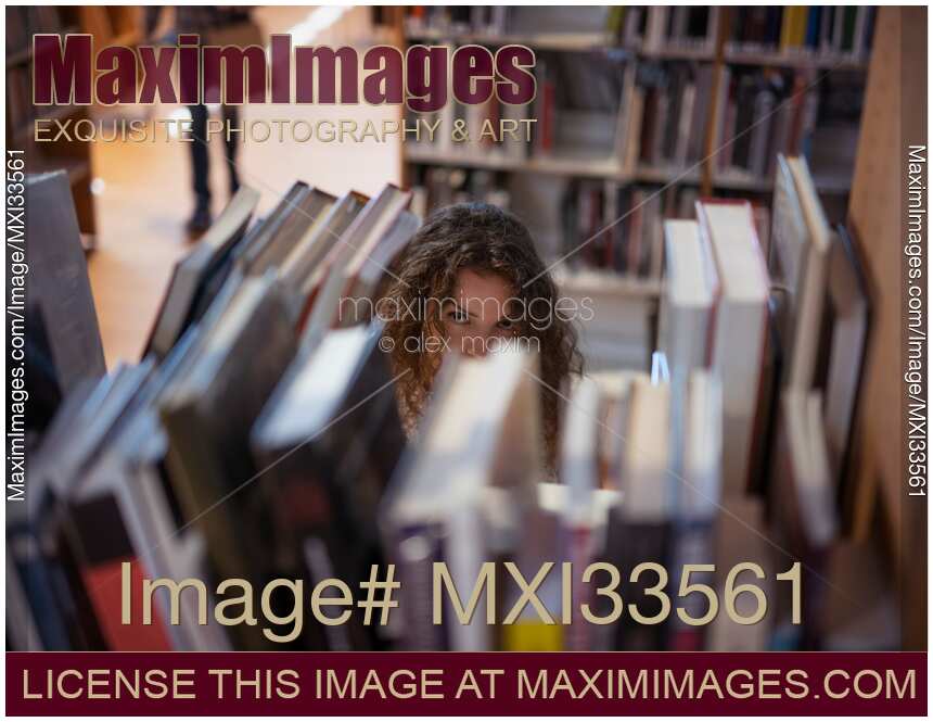 Young woman, college student, looking from behind a book shelf in a library
