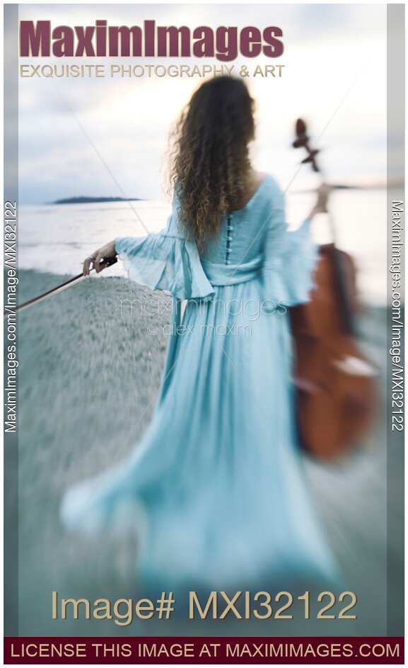 Young woman in a long dress walking towards the ocean shore with a cello in an atmospheric scenery