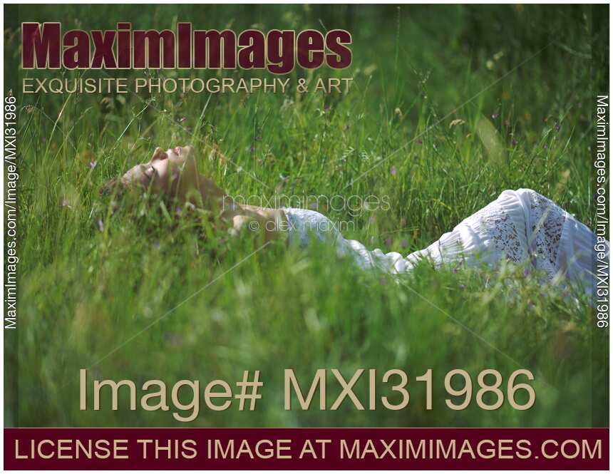 Young woman in a summer dress lying in green grassy field facing the sunlight in summer nature scenery
