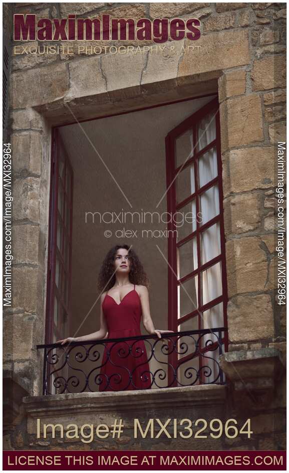 Young woman in red dress standing in an open balcony window of an old French house looking outside