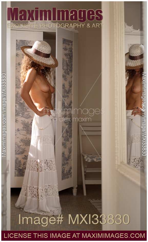 Young woman in sun hat and white dress standing topless by wardrobe reflecting in mirror in sunlit French vintage room