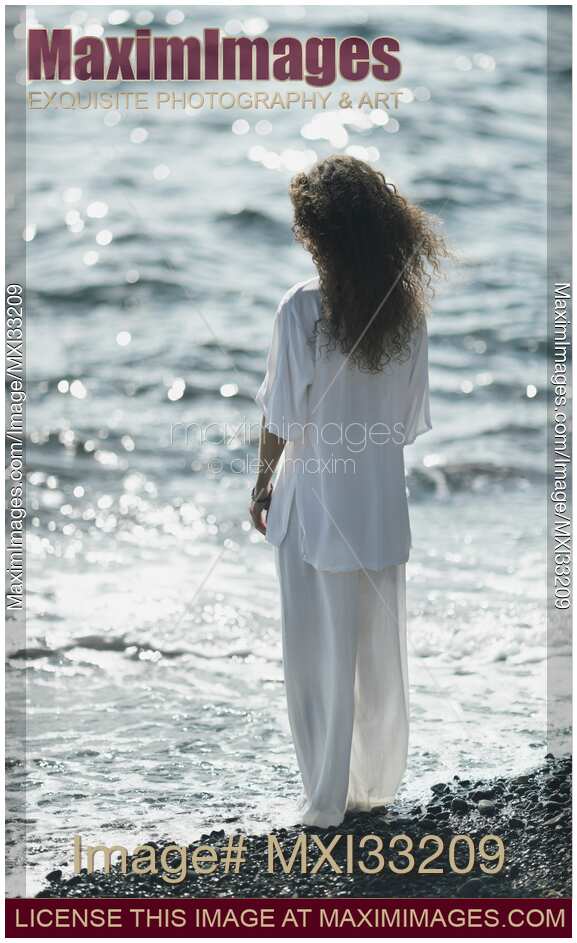 Young woman in white summer clothes standing on a pebble ocean shore with cool water washing over her bare feet