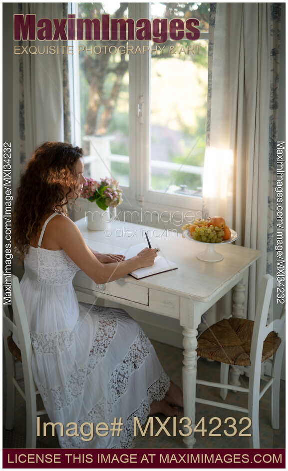 Young woman in white summer dress writing in a diary sitting at a table by a window in country house