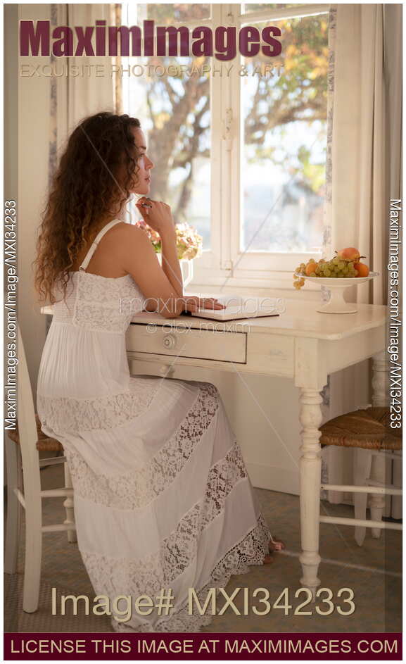Young woman writing in a diary sitting at a table by a window with a thoughtful expression