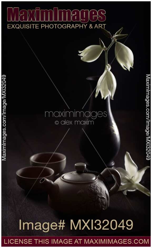 Zen still life of a clay teapot with tea cups and flowers in antique vase on black