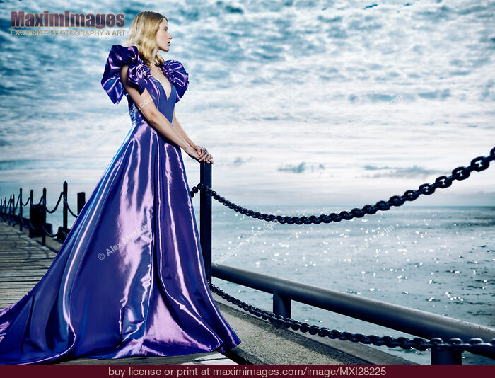 Stock photo of Woman wearing beautiful long blue dress standing at waterfront Buy commercial use license at MaximImages