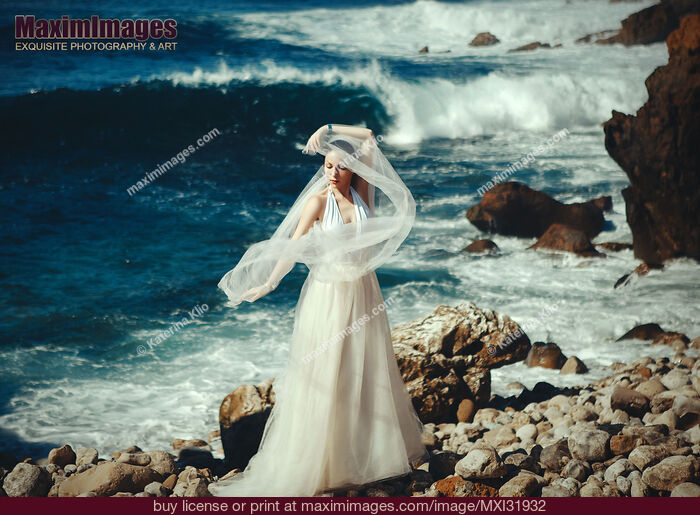 Stock photo of Woman in a white wedding dress standing on a rocky sea shore with a veil in the wind Buy commercial use license at MaximImages