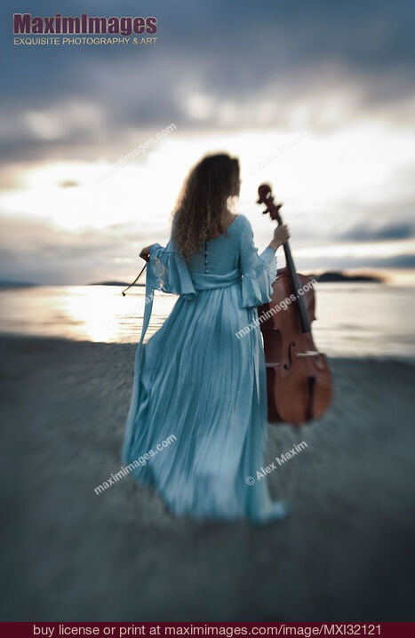 Stock photo of Atmospheric portrait of a woman in a vintage dress standing with a cello on an ocean shore in the wind in dramatic sunset Buy commercial use license at MaximImages