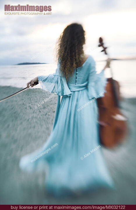 Stock photo of Young woman in a long dress walking towards the ocean shore with a cello in an atmospheric scenery Buy commercial use license at MaximImages