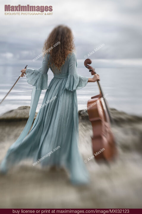 Stock photo of Woman in a long vintage dress standing gracefully by the ocean shore with a cello and a bow Buy commercial use license at MaximImages