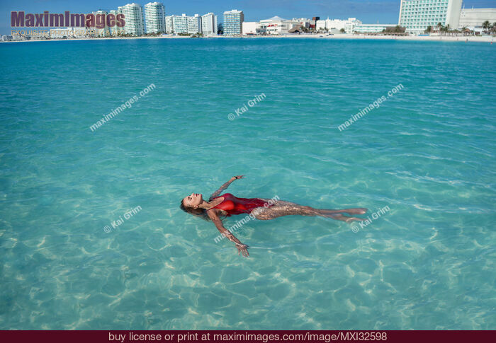 Stock photo of Woman in red swimsuit lying in clear blue water of Caribbean Sea with Cancun city skyline in the background Buy commercial use license at MaximImages
