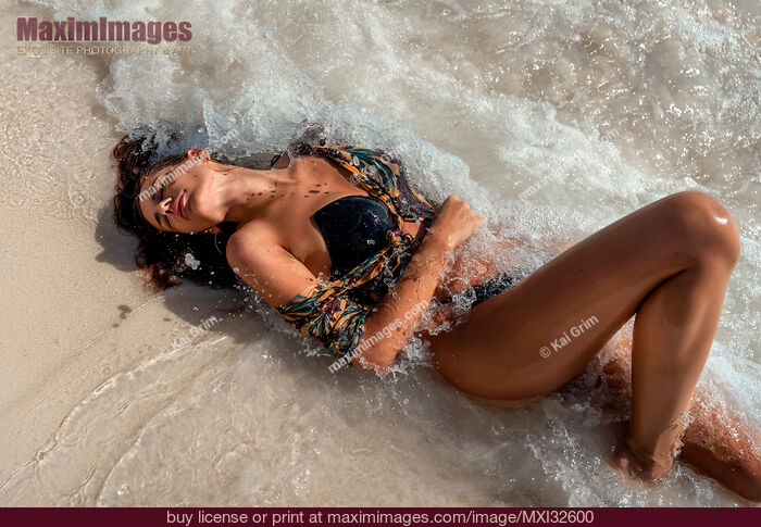 Stock photo of Sexy woman in swimsuit lying on a sandy shore with splashing waves of the Caribbean Sea hitting her body Buy commercial use license at MaximImages