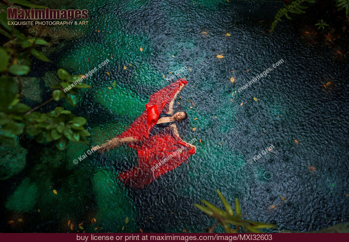 Stock photo of Woman in swimsuit and red dress lying on blue water in the rain Beautiful aerial fine art photograph Buy commercial use license at MaximImages