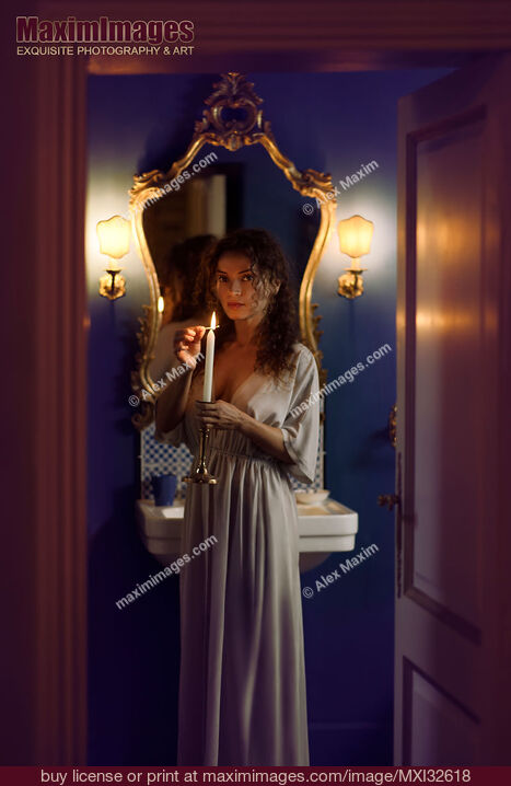 Stock photo of Beautiful woman in evening gown lighting a candle in her hand with an expressive mysterious look in bathroom settings Buy commercial use license at MaximImages