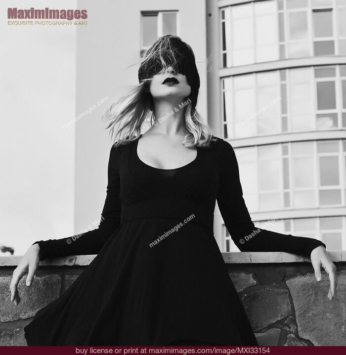 Stock photo of Woman in black dress standing blindfolded on a balcony in outdoor city scenery Buy commercial use license at MaximImages