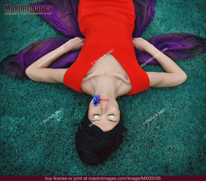 Stock photo of Surreal portrait of an extravagant lady in red dress lying on blue grass with a flower in her mouth Buy commercial use license at MaximImages
