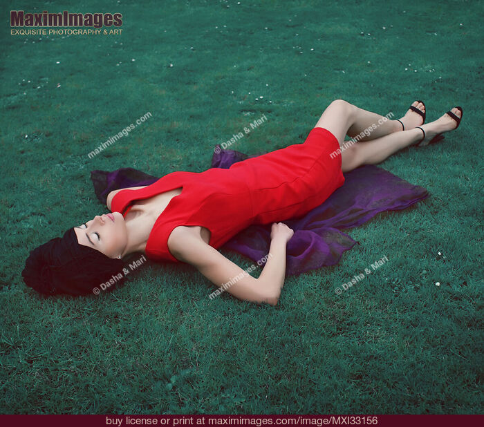 Stock photo of Surreal fashion portrait of extravagant woman in bright red dress lying on blue green grass Buy commercial use license at MaximImages