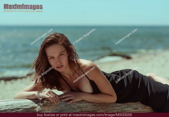Stock photo of Sensual portrait of a young woman in black dress posing on a driftwood at the beach Buy commercial use license at MaximImages