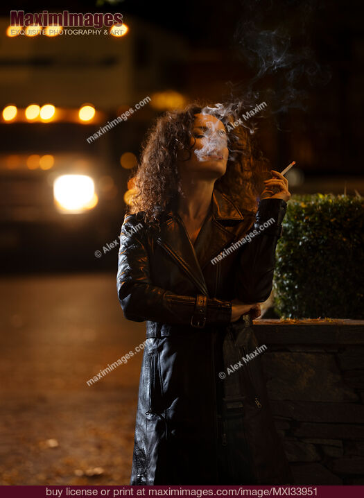 Stock photo of Woman in black coat smoking a cigarette standing on a night city street exhaling smoke Buy commercial use license at MaximImages