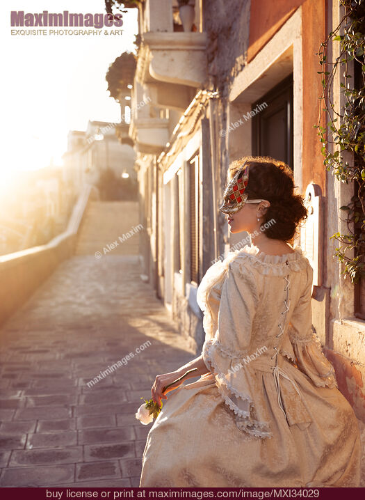 Stock photo of Beautiful lady in vintage Italian carnival dress and mask walking along sunlit historic street in Venice Buy commercial use license at MaximImages