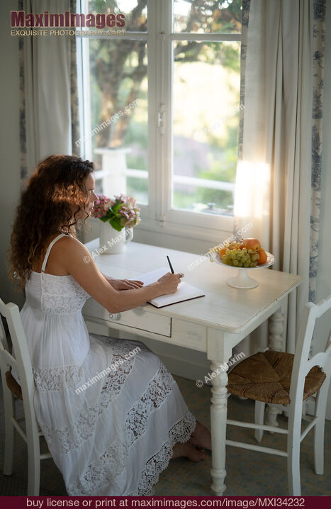 Stock photo of Young woman in white summer dress writing in a diary sitting at a table by a window in country house Buy commercial use license at MaximImages