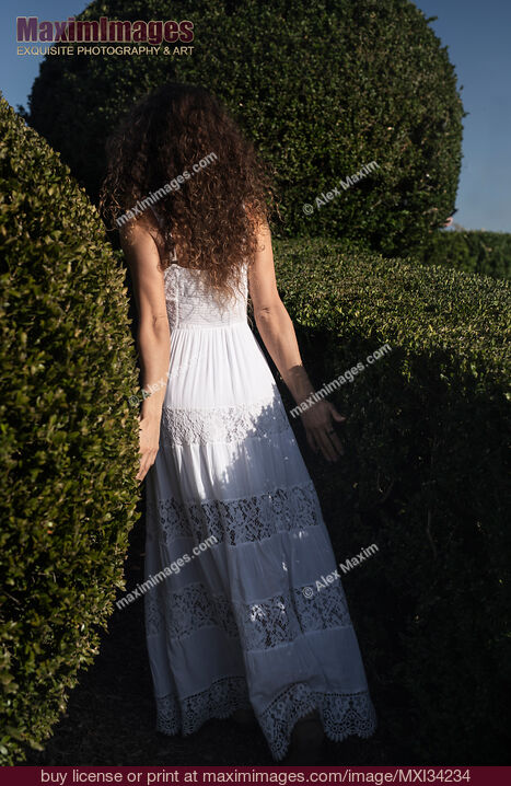 Stock photo of Young lady in white summer dress walking into a green garden disappearing behind the bushes Buy commercial use license at MaximImages