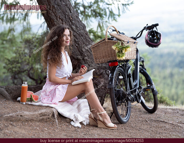 Stock photo of Young pretty woman sitting on a blanket with book and bike enjoying a little picnic in summer countryside Buy commercial use license at MaximImages