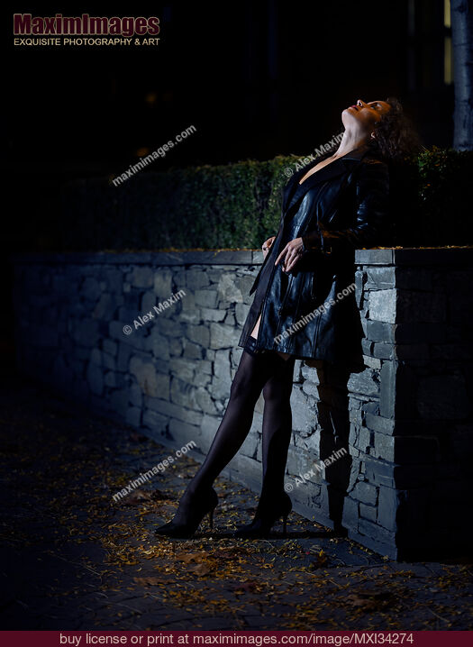 Stock photo of Sexy woman dressed in black leather coat and black stockings standing on city street looking at night sky Buy commercial use license at MaximImages