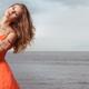 Young woman in orange dress standing on a shore... (thumbnail)