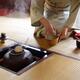 Woman blending matcha tea with a whisk by a steaming iron kettle in a... (thumbnail)